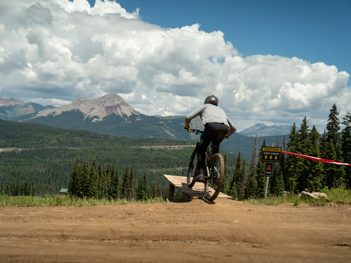 An adventurous mountain biker launches off a wooden ramp, with breathtaking mountains towering in the background. The image radiates energy, but the vast, open space and spectacular views also invite exploration and shared outdoor experiences.