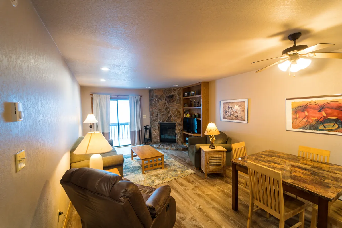A spacious, inviting living area with a central fireplace, a coffee table perfect for board games or warm drinks, and a dining table nearby. Large windows with curtains let in ample natural light, enhancing the sense of warmth and relaxation.