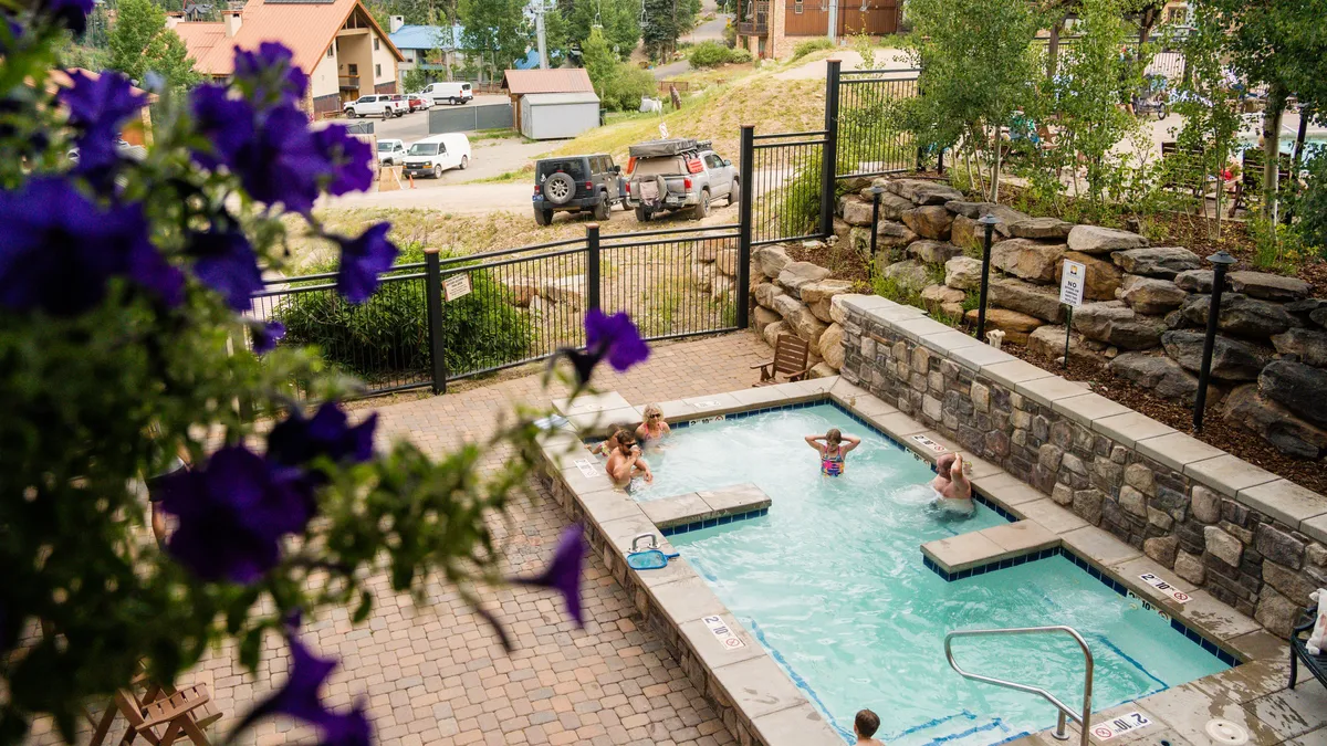 An outdoor hot tub where children and adults are enjoying the water, surrounded by lush greenery, stone walls, and vibrant flowers. The joyful expressions and playful atmosphere highlight family-friendly experiences.