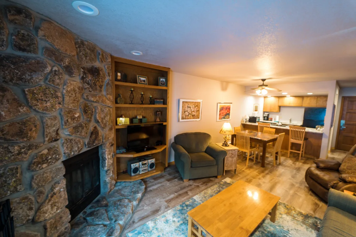 The image showcases a cozy interior featuring a striking stone fireplace, a warm wooden dining table set for gatherings, plush seating perfect for relaxing, and decorative shelves adding homely touches. The ambiance is inviting and ideal for unwinding after a day in the mountains.