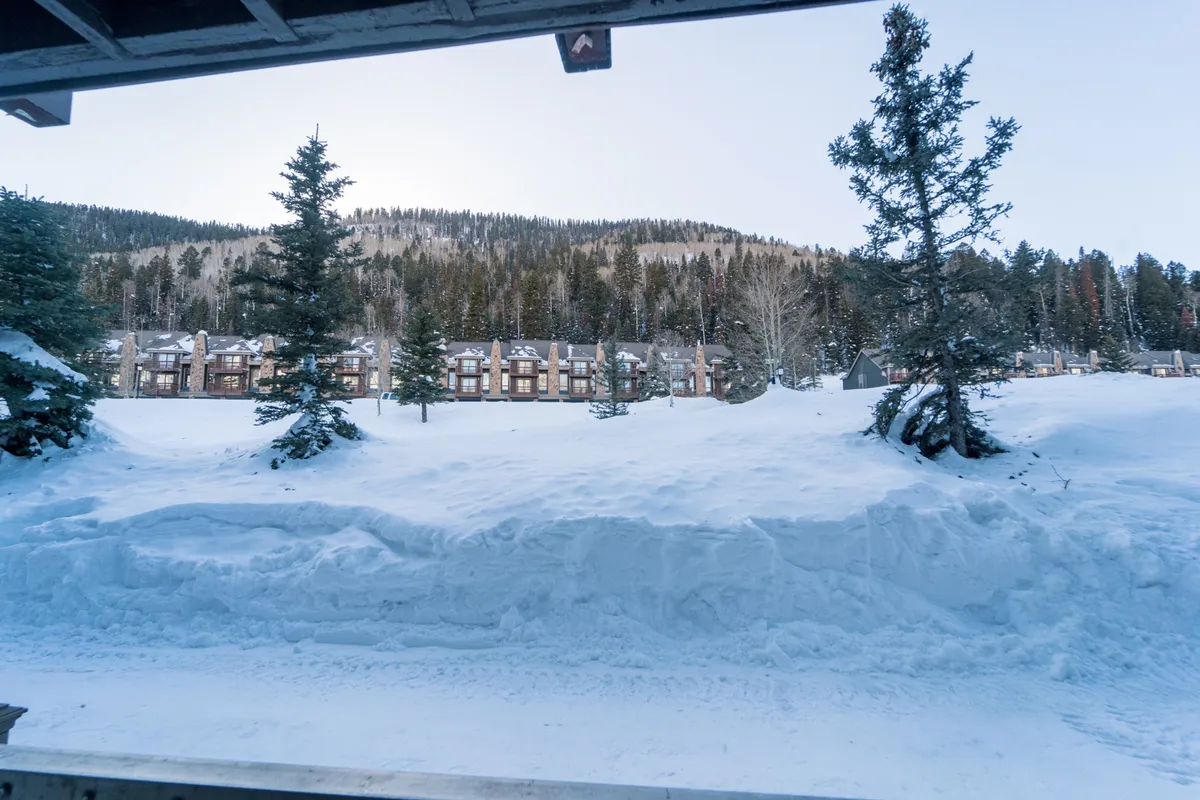 A pristine snow-covered landscape frames a row of cozy townhouses, with towering pine trees and a distant mountain range adding to the sense of winter wonder. The scene suggests peaceful evenings and the embrace of the mountain environment, away from the bustle of the resort.