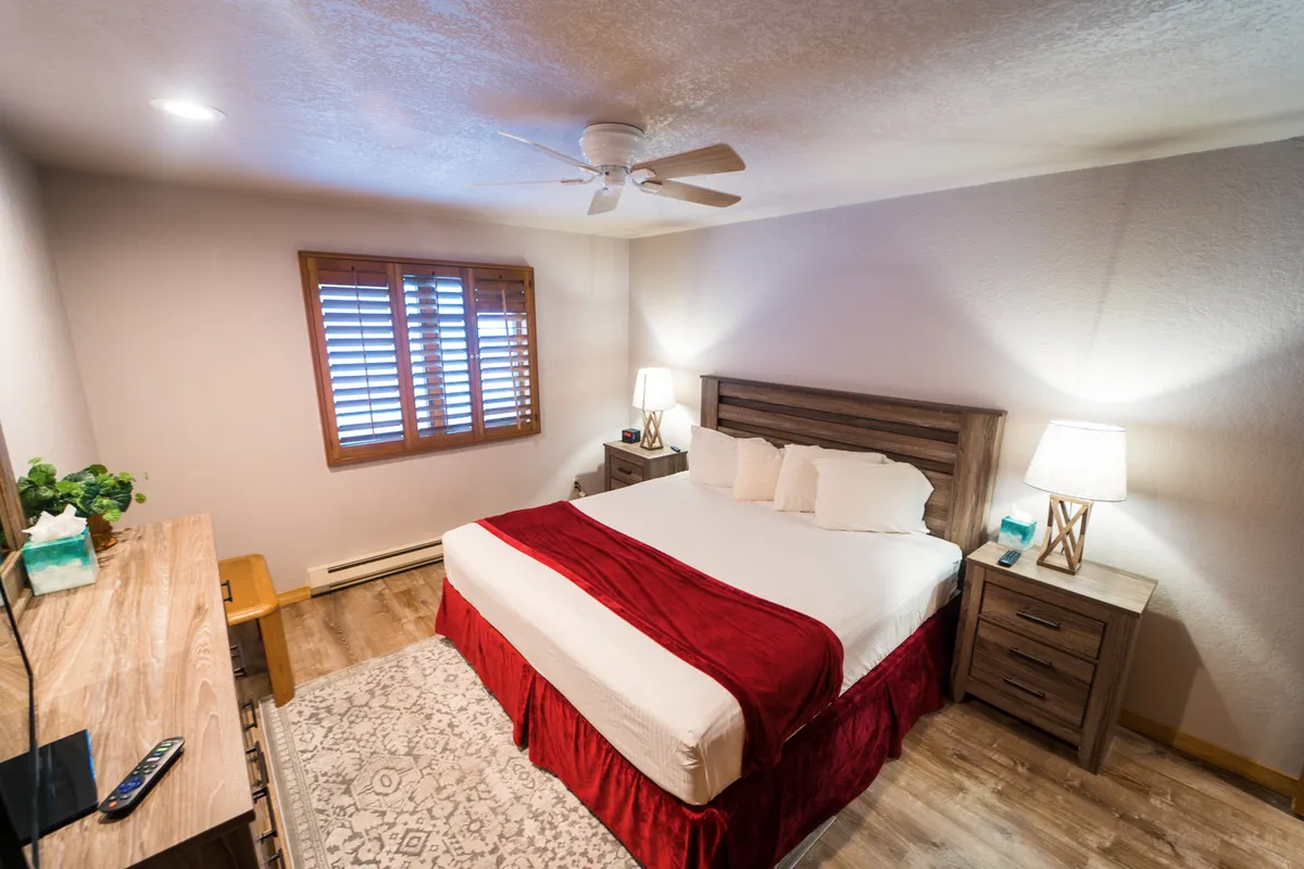 A cozy bedroom featuring a comfortable bed with a red accent throw, matching nightstands with softly glowing lamps, and a shuttered window that lets in gentle natural light. The space feels tailored for relaxation, with thoughtfully arranged details that enhance the serene atmosphere.