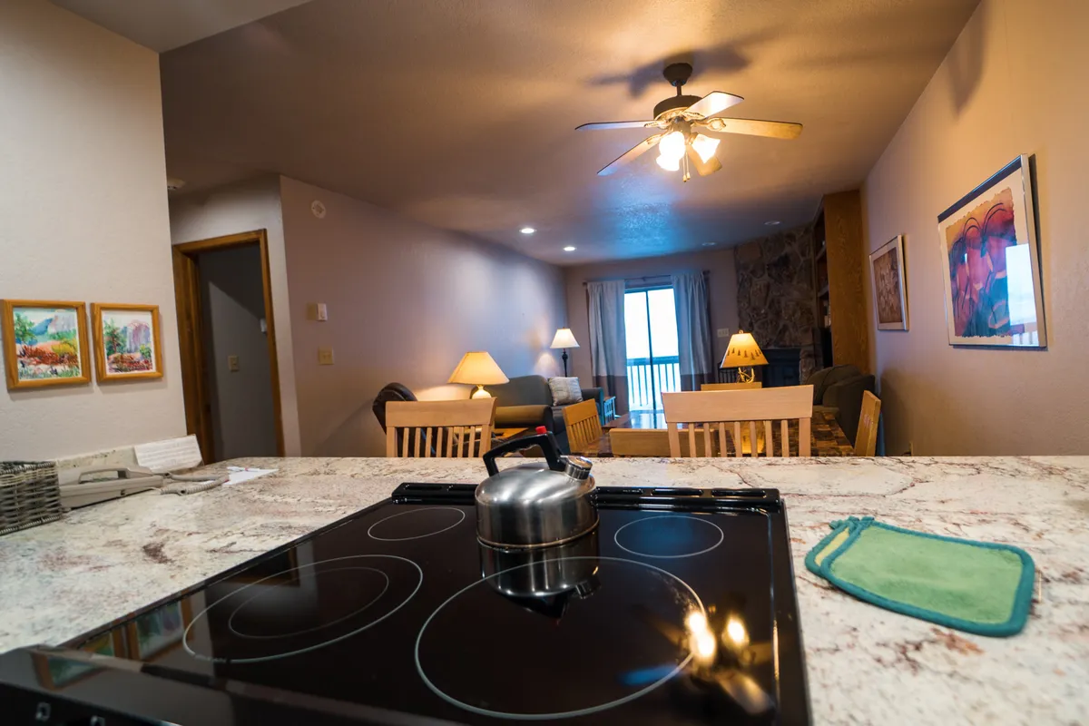 A wide view of the open kitchen and dining area, featuring a kitchen island with a stainless steel kettle, a cozy living area, and a dining table. The scene also hints at a balcony view, expanding the sense of space and comfort.