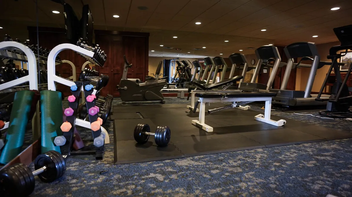 A bright, well-equipped fitness center featuring treadmills, dumbbells, an exercise bench, and weightlifting equipment. The space is open and inviting, encouraging guests to enjoy a workout or join community fitness activities.