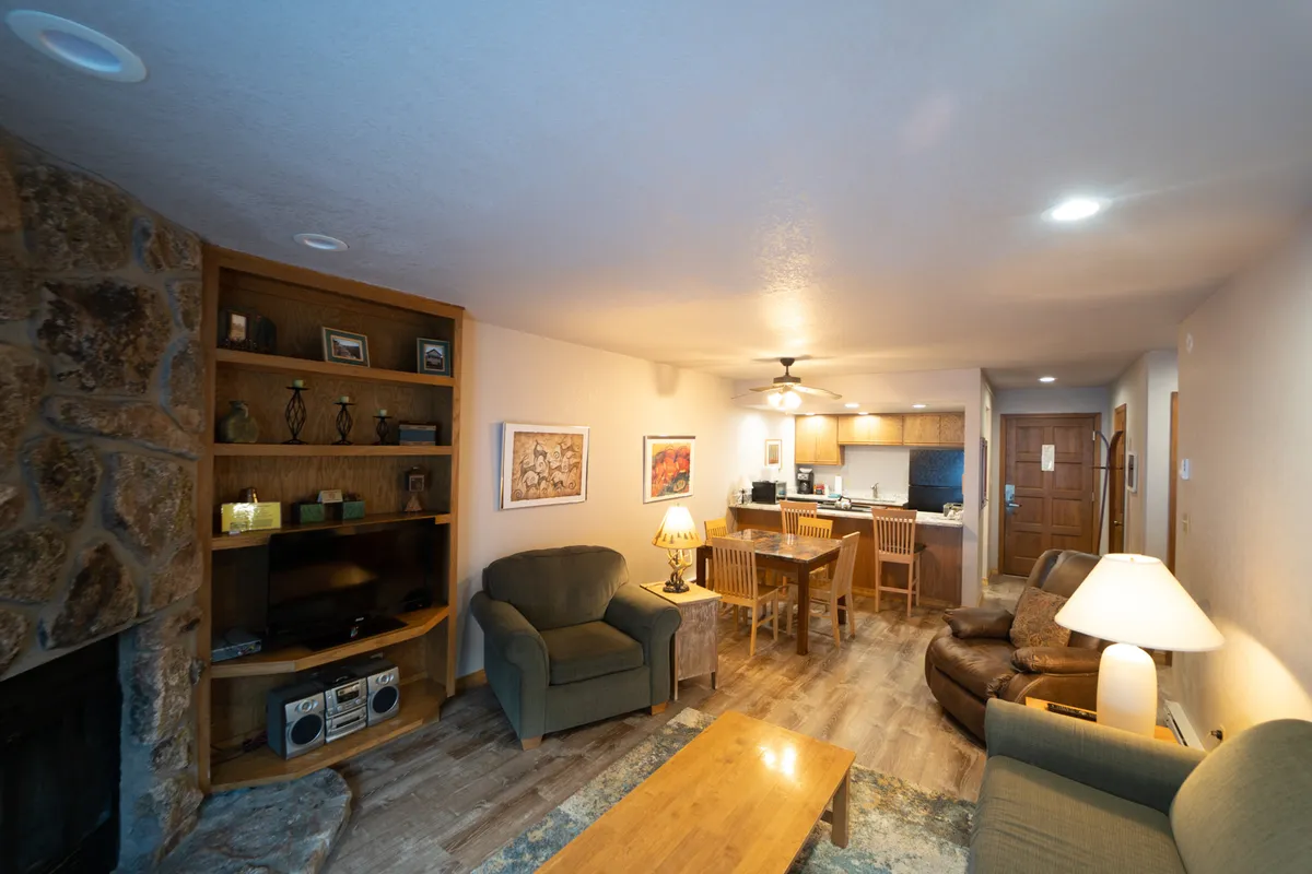 A welcoming living room featuring a stone fireplace as the focal point, surrounded by a wooden shelving unit, a cozy dining table, and comfortable seating arranged to create an intimate conversation area. Soft mountain light bathes the space, highlighting the inviting textures and warm tones.