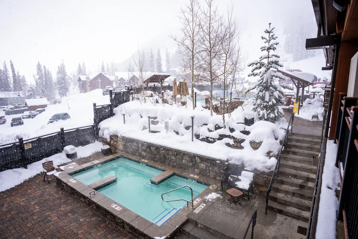 This image features an inviting hot tub set against a snow-covered landscape, with wooden structures and trees surrounding the space. Guests are seen relaxing in the warm water while socializing near the fire pit, creating a cozy and luxurious winter atmosphere.