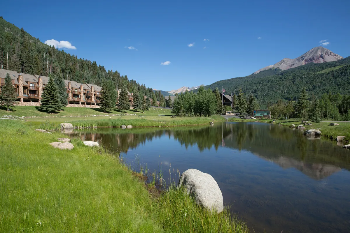 A tranquil pond sits at the heart of lush green grounds, reflecting the brilliant mountain backdrop. Cozy wooden lodge buildings nestle at the edge of the scene, suggesting a peaceful, nature-focused setting ideal for couples seeking serenity.
