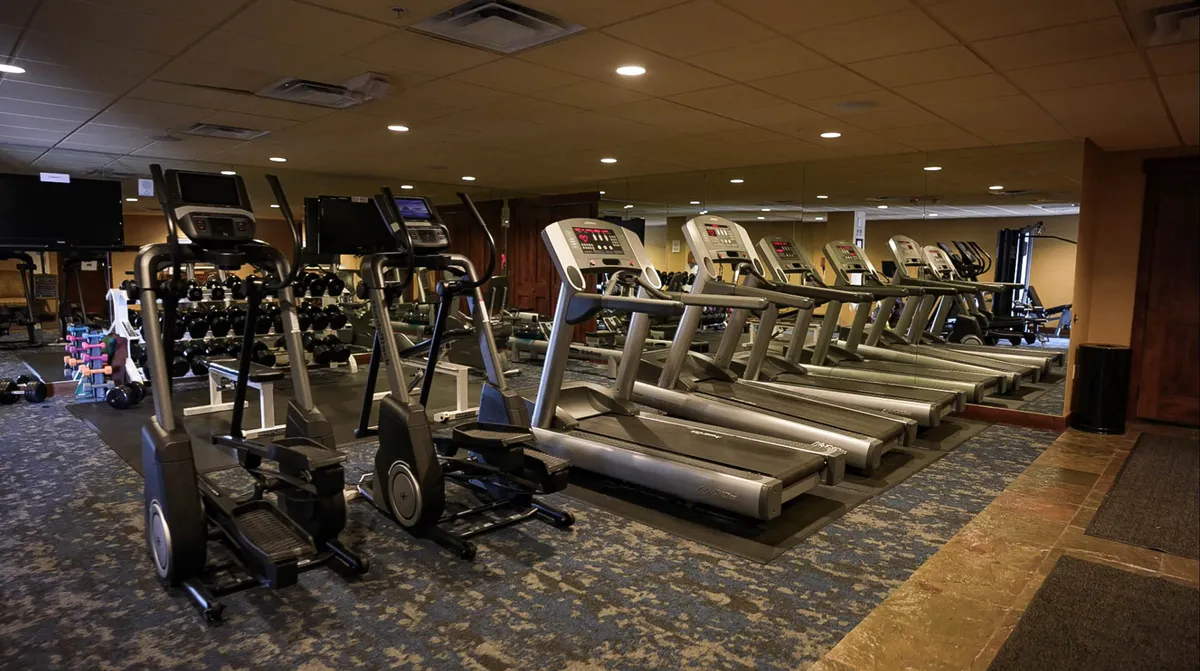 A bright, modern fitness center equipped with treadmills, elliptical machines, dumbbells, and exercise mats, all facing a mirrored wall. The space is designed for invigorating workouts and communal fitness activities.
