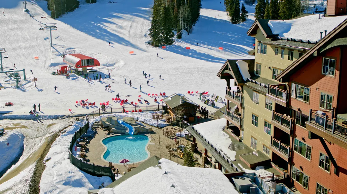 A vibrant exterior winter scene featuring ski slopes with skiers, an outdoor pool surrounded by snow, a ski lift in operation, and a cozy lodge building. The sun is setting, casting a festive glow over the slopes and gathering areas.