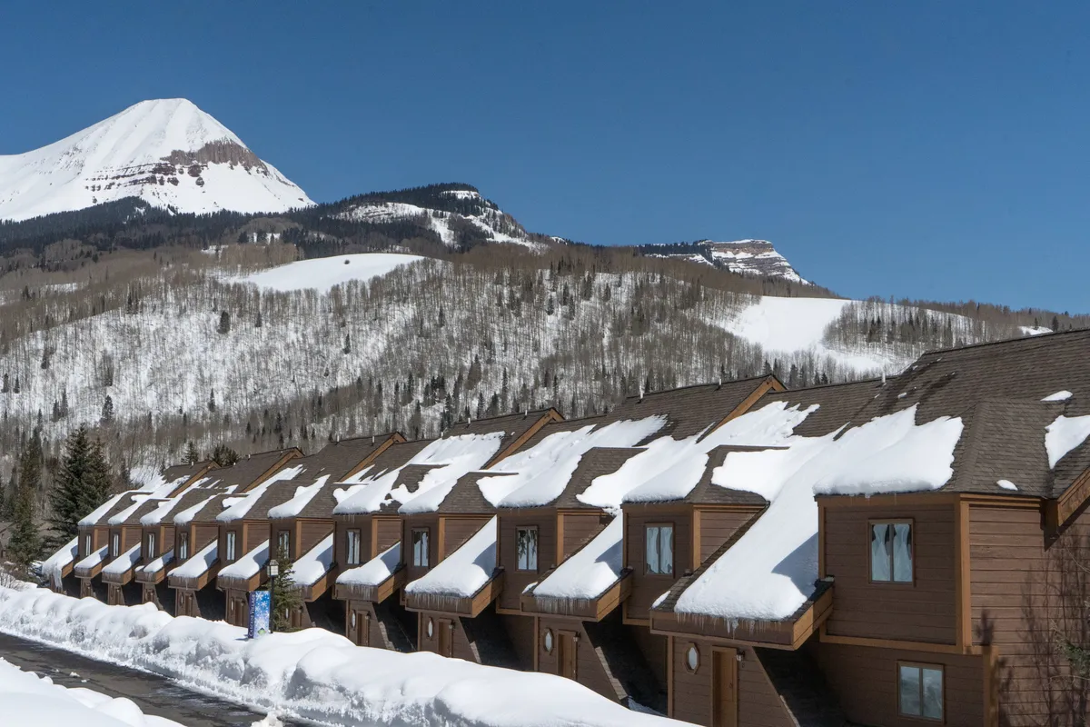 A tranquil winter exterior with snow-covered rooftops against a dramatic mountain backdrop and a clear blue sky. The scene evokes relaxation after winter activities, family fun in the snow, and the serene beauty of the mountain setting.